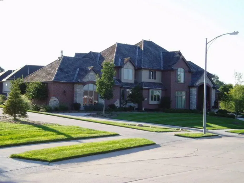 Large red brick suburban house with dark gray shingles, a manicured lawn, and a streetlamp in the foreground.