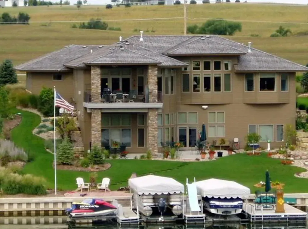 A large tan house with stone accents, a multi-level deck, and a green lawn overlooking a dock with boats and a jet ski.