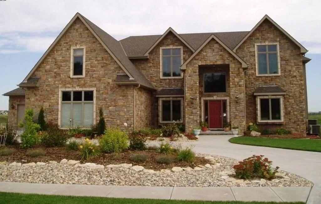 Two-story house with tan stone exterior, brown roof, and red front door, featuring a concrete driveway and landscaping.