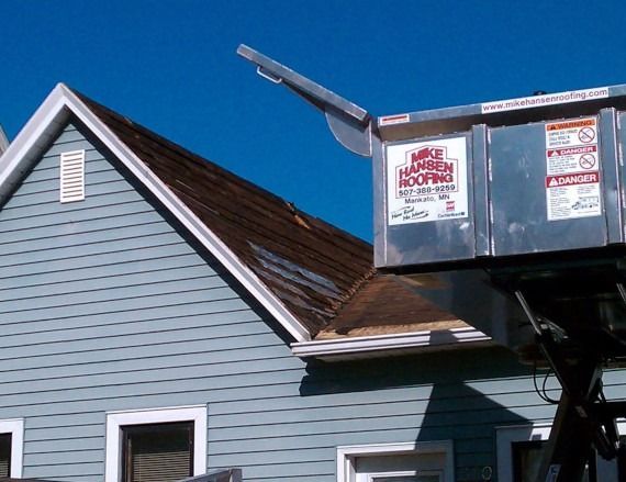Blue house roof being repaired with a raised dumpster beside it under a clear sky