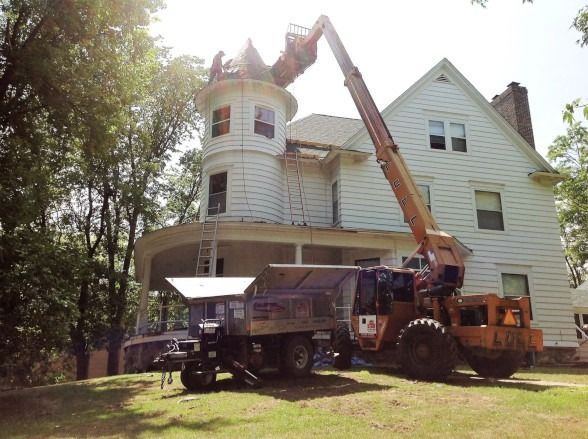 Construction equipment lifting a dump truck beside a white house with a curved tower porch