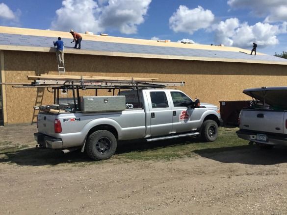 Workers on a roof above parked trucks beside a tan building on a sunny day