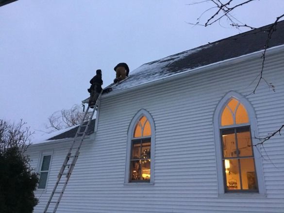 Two people on a ladder work on a snowy roof above lit arched windows at dusk