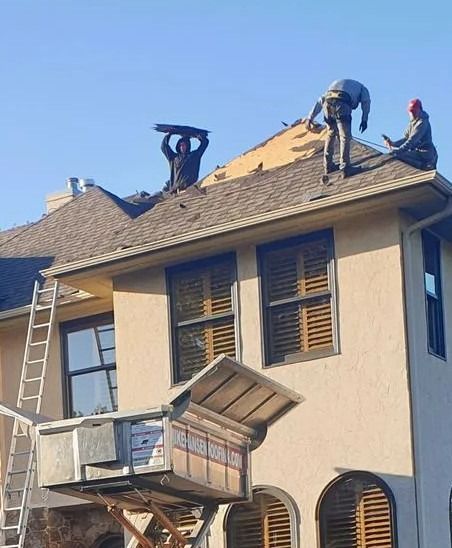 Workers repairing a roof on a beige house, with ladders and a raised work platform.