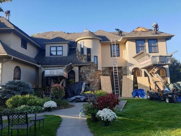 Large two-story stucco house with stone accents, front lawn, and outdoor stairs in a sunny hillside setting