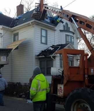 Workers and a lift truck beside a house with a damaged roof during repairs