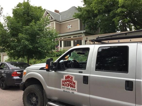 White utility truck parked on a street in front of a brick building with trees and another car nearby