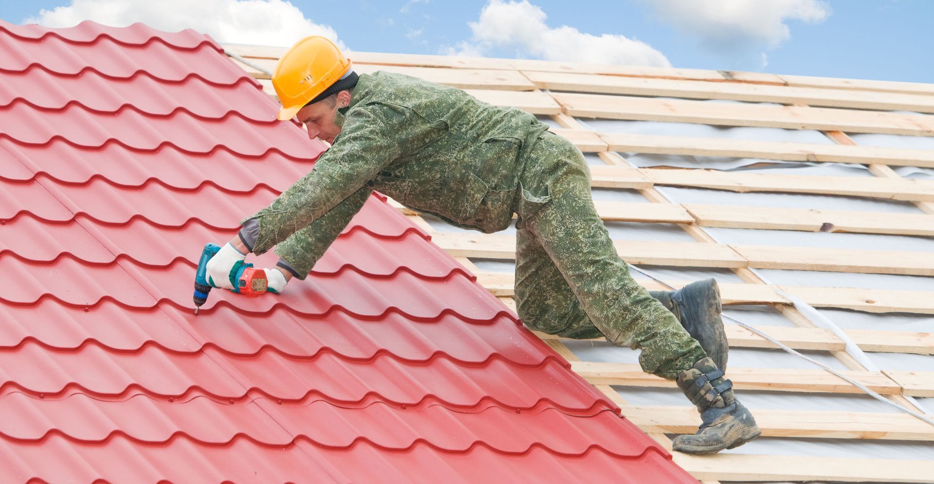 Worker installing red roof tiles on a rooftop, wearing a yellow hard hat and green work clothes