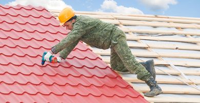 Worker installing red roof tiles on a rooftop, wearing a yellow hard hat and green work clothes