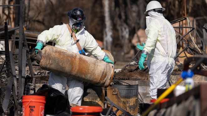 Two people in hazmat suits carrying a large, cylindrical object amidst debris; outdoors.