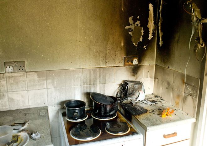 Kitchen interior severely damaged by fire; burnt appliances and walls.