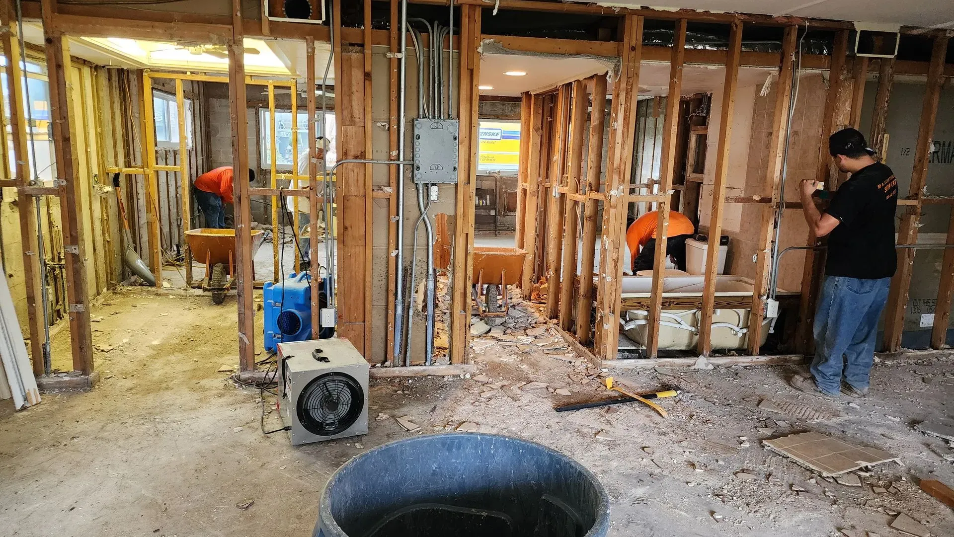 Interior demolition of a home; workers, exposed framing, rubble, fan, and trash can.