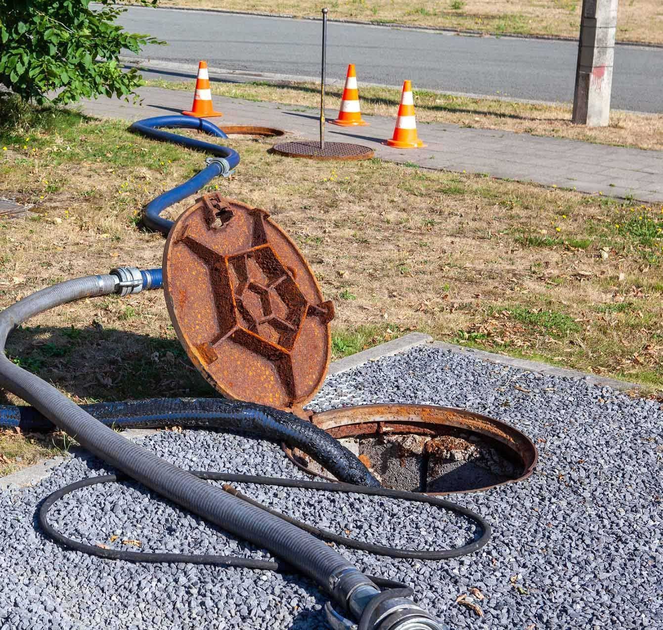 Sewer lid open, with hoses and traffic cones, near a sidewalk and road, indicating maintenance in progress.