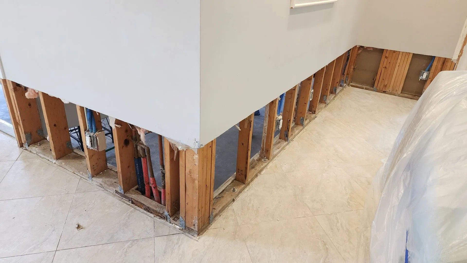 Interior wall gutted, revealing wooden studs, pipes, and electrical wiring. White wall meets corner. Floor tiles present.
