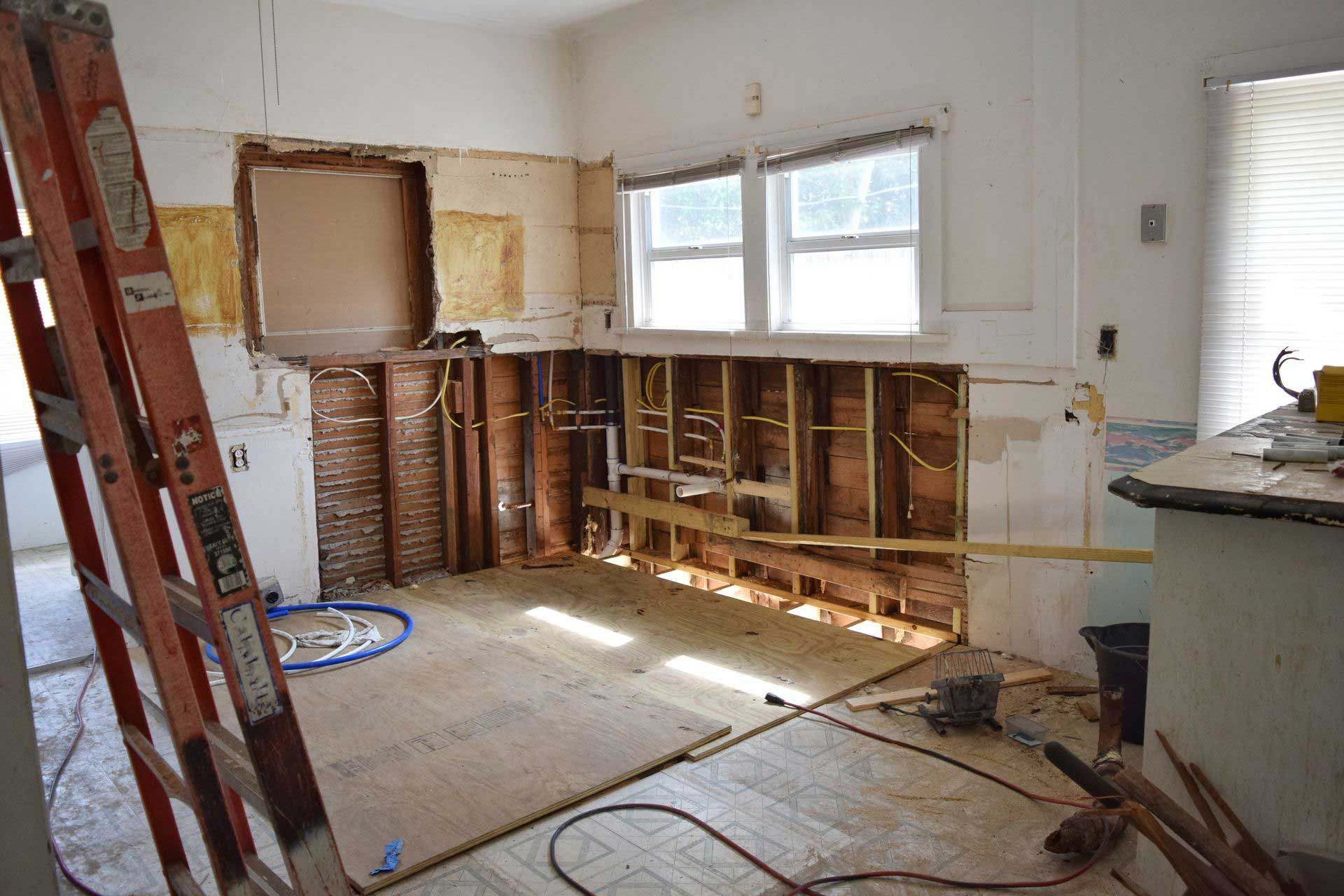 Kitchen during renovation: walls stripped to studs, ladder leaning against the wall, wood flooring, window in the background.