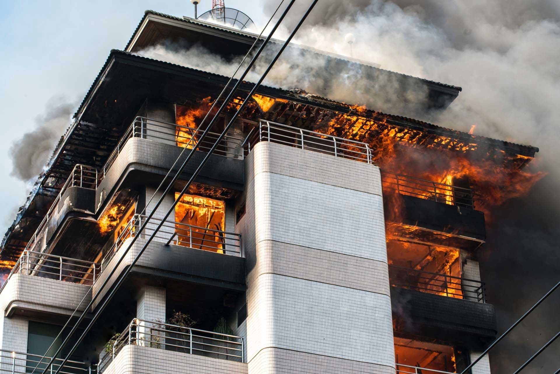 Burning building; flames and smoke engulf several floors, power lines visible, bright sky.