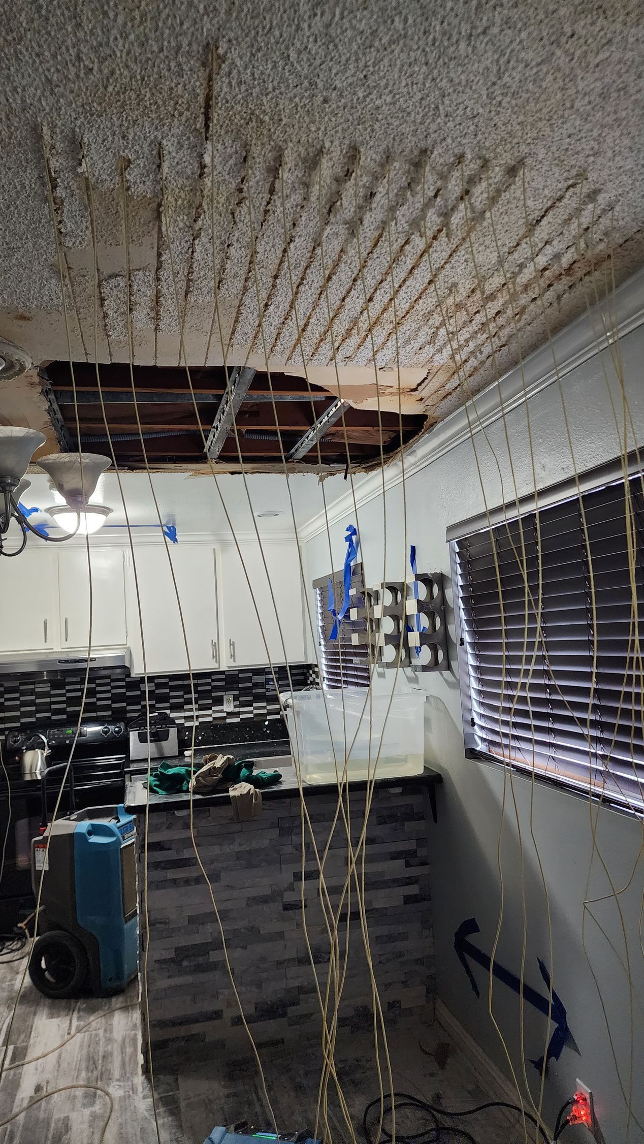 Water-damaged room. Ceiling partially collapsed, blue equipment on floor, window with blinds, dark flooring.