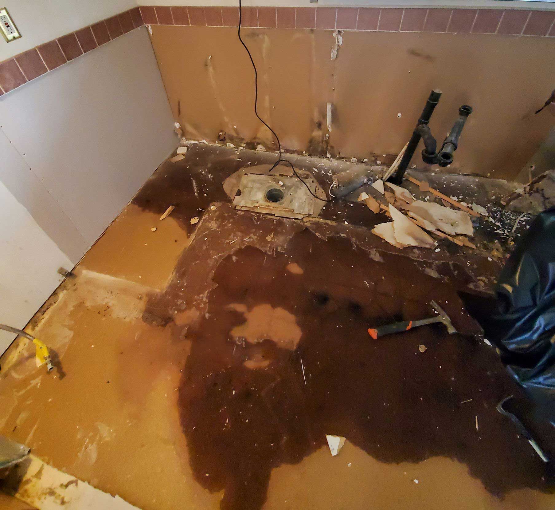 Bathroom renovation in progress, damaged floor with plumbing visible. Walls stripped of tile, debris scattered.