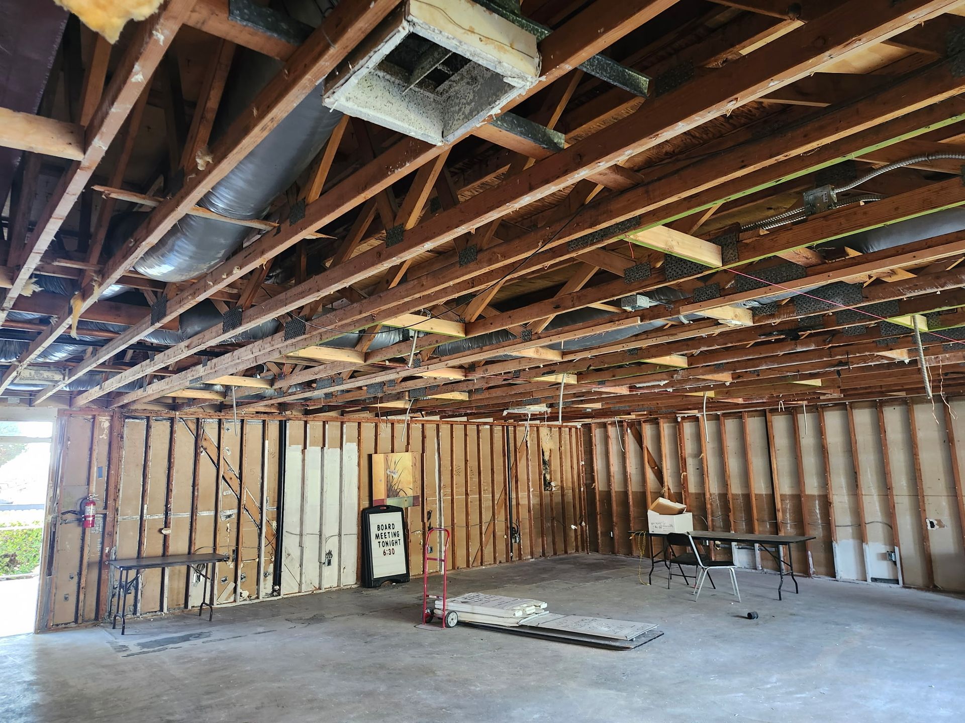 Interior of a room under construction, with exposed wooden beams, studs, and ductwork. Cement floor.
