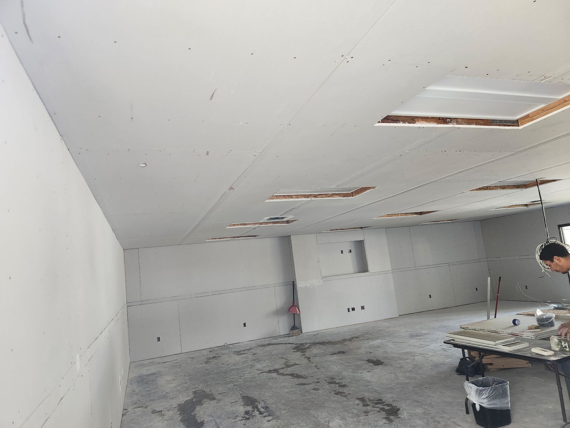 Interior construction with drywall on walls and ceiling. Exposed holes. Man working near table.