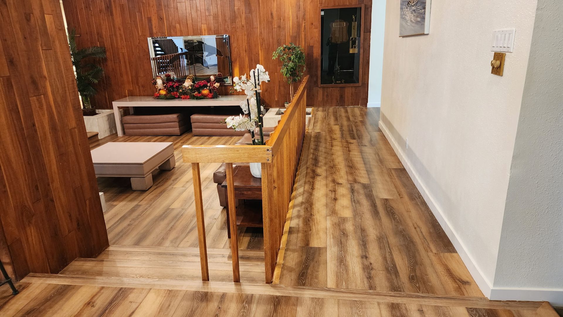 Wooden-paneled living room with a TV, plants, and staircase leading to a hallway with wood-look flooring.