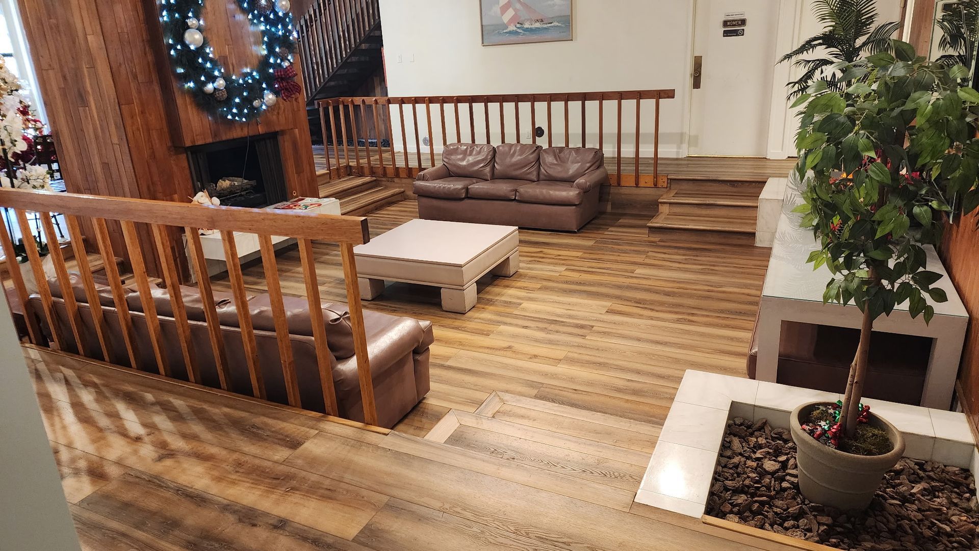 Lounge area with brown leather sofas, coffee table, fireplace, and potted tree on a wood-plank floor.