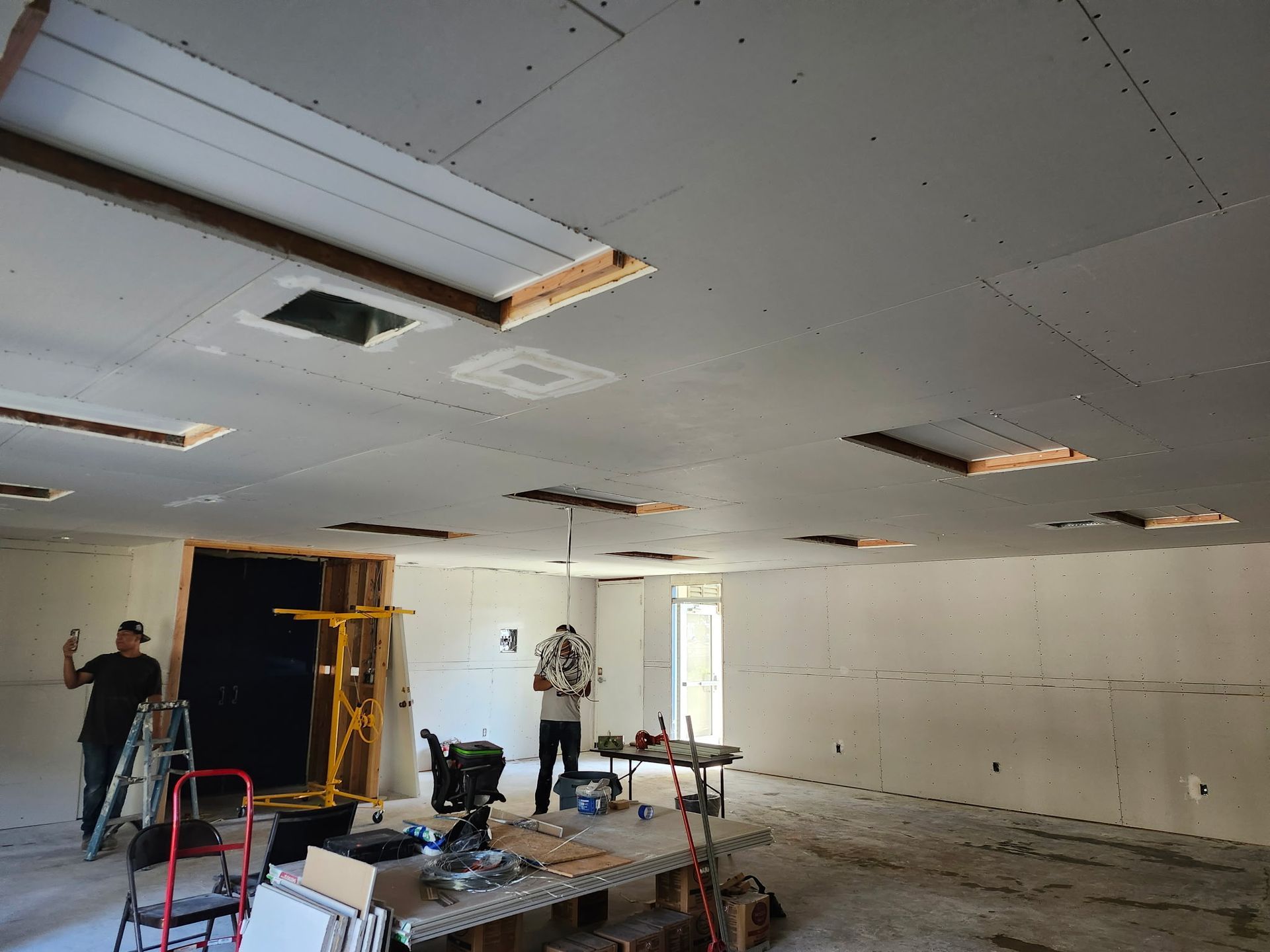 Workers install drywall on a ceiling with multiple recessed light openings. Room is under construction.