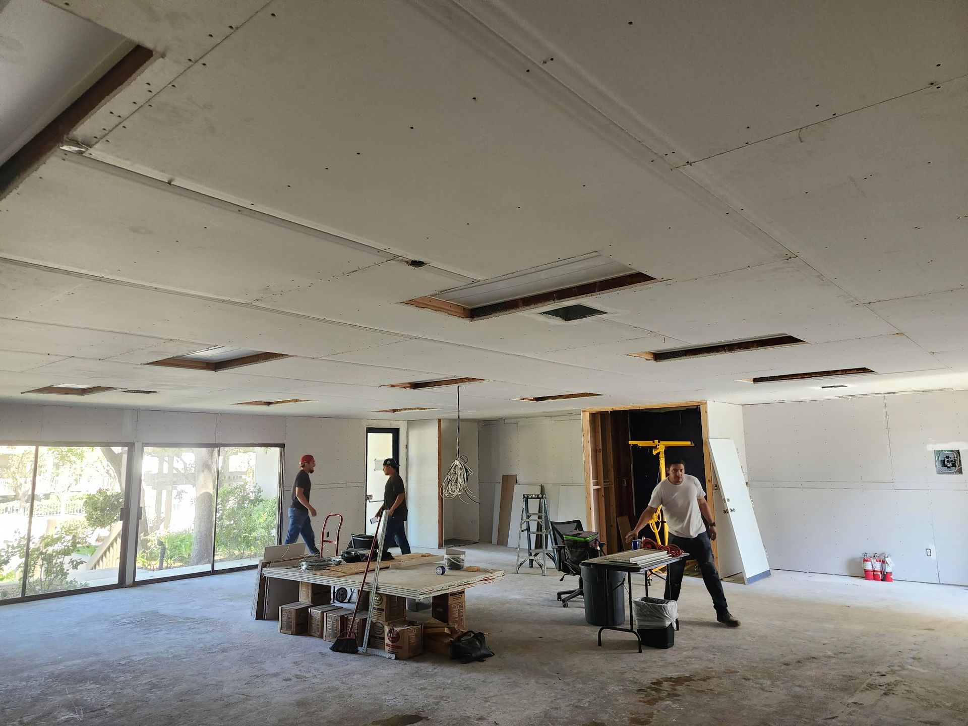 Construction crew working on ceiling and walls in a room with large windows.