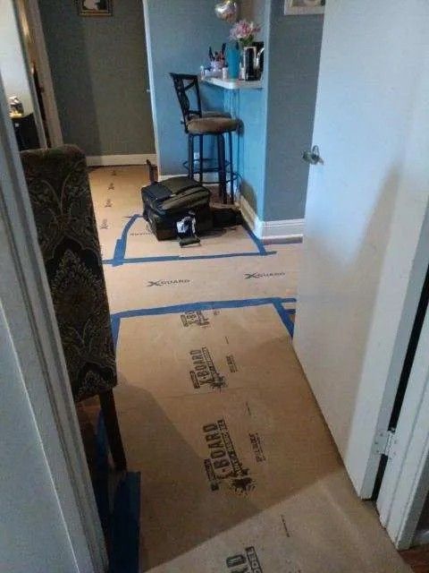Flooring protection in progress. Cardboard covers hardwood floors, taped off in room with chair, bar stool, and blue walls.