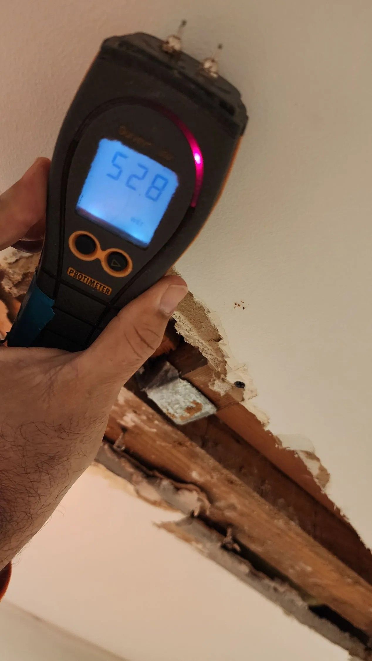 A moisture meter reads 528 on a damaged ceiling. The ceiling is open, revealing wooden beams. A hand holds the meter.