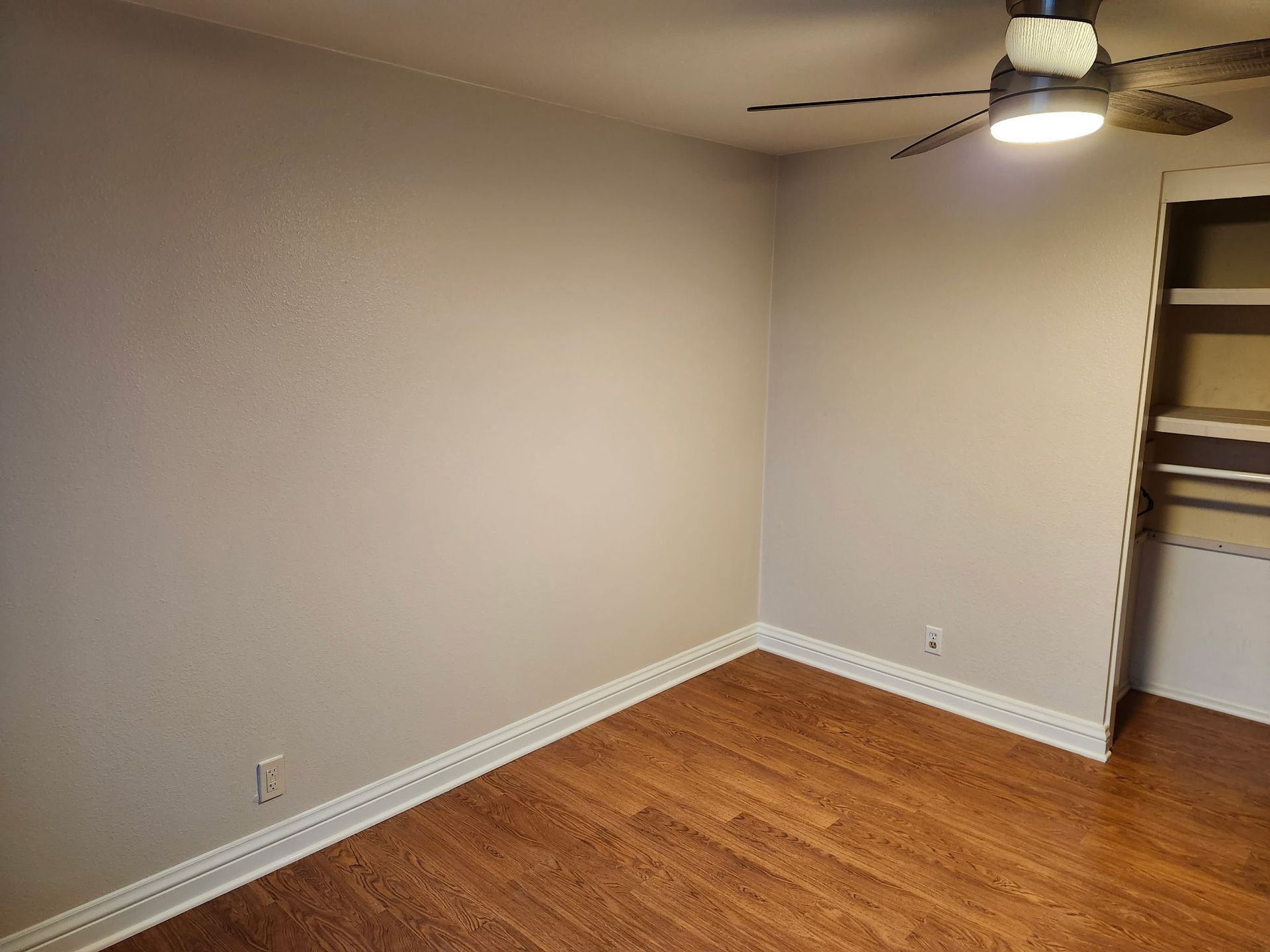 Empty room with hardwood floors, beige walls, and a ceiling fan. A built-in shelf is visible.
