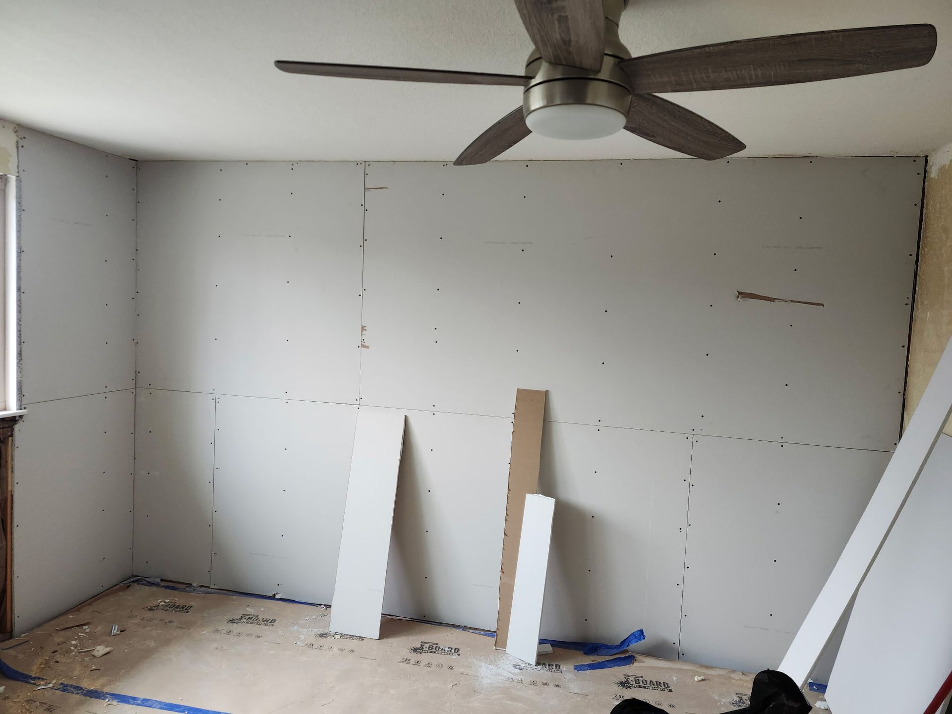 Room with drywall installed, unfinished. Ceiling fan visible. Wood pieces on floor.