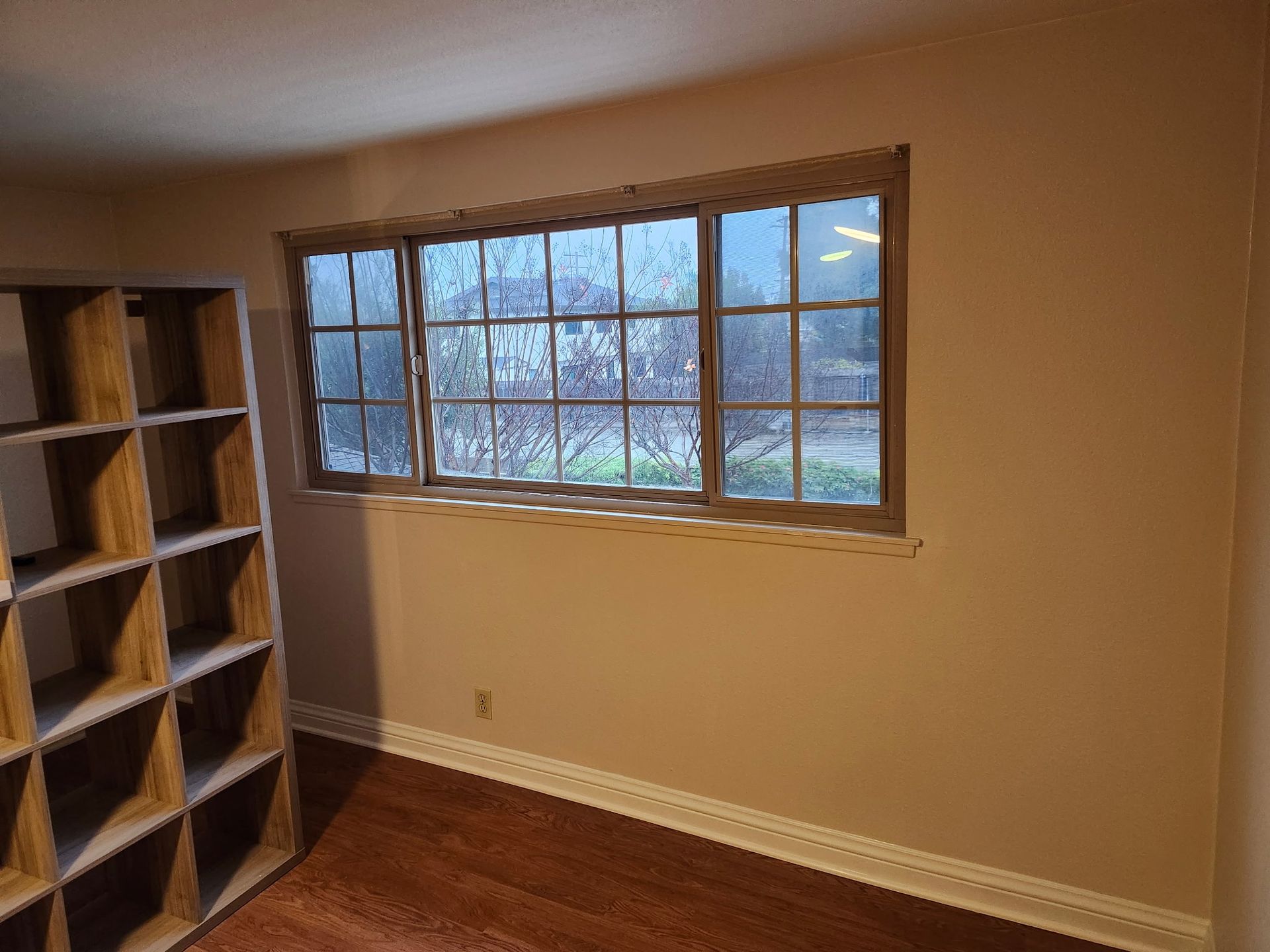Empty room with a window, shelves, and hardwood floor. The walls are beige, and the window has a light brown frame.