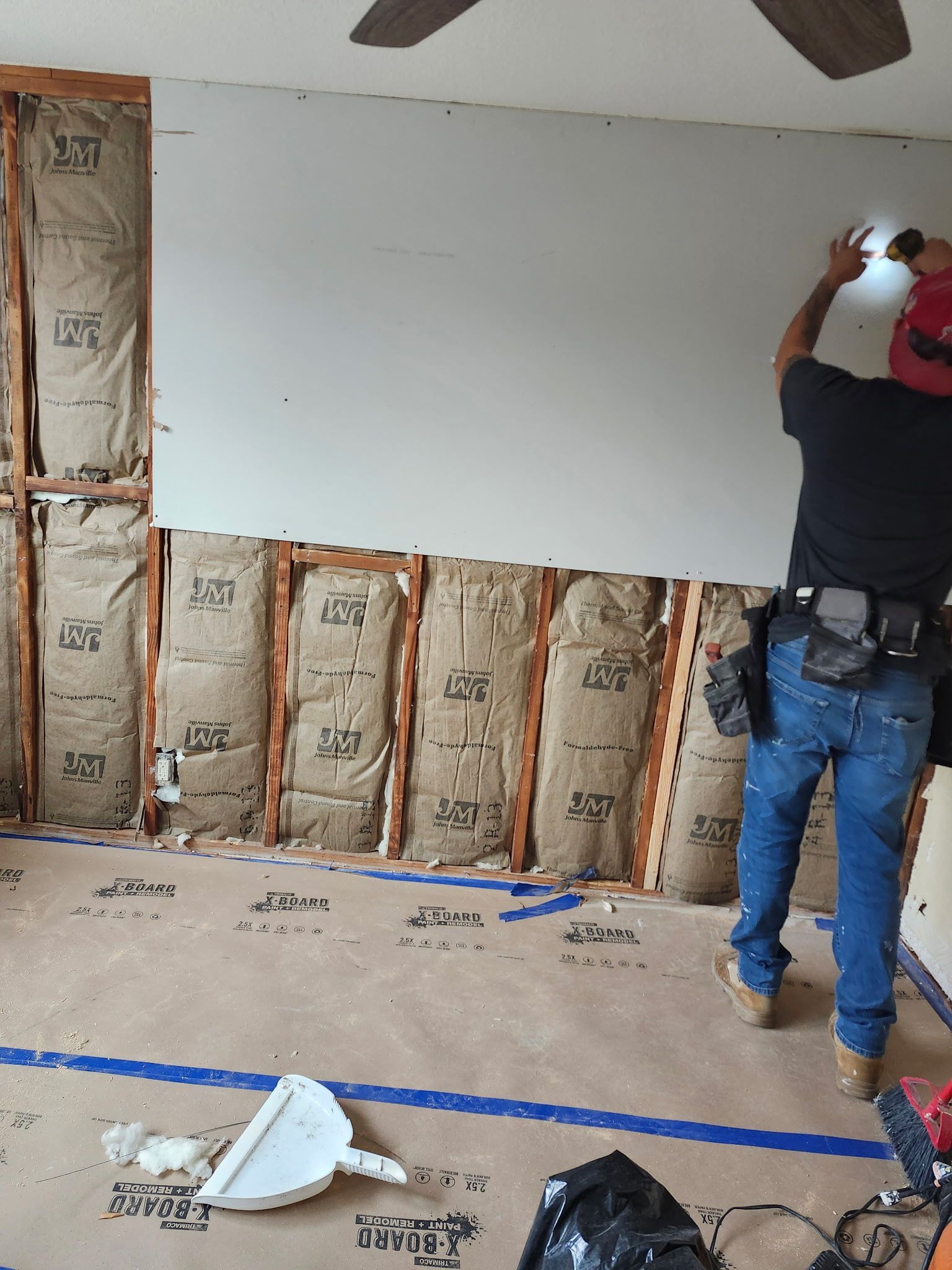 A person installs drywall on a wall with exposed insulation. A toolbelt is visible. Indoors.