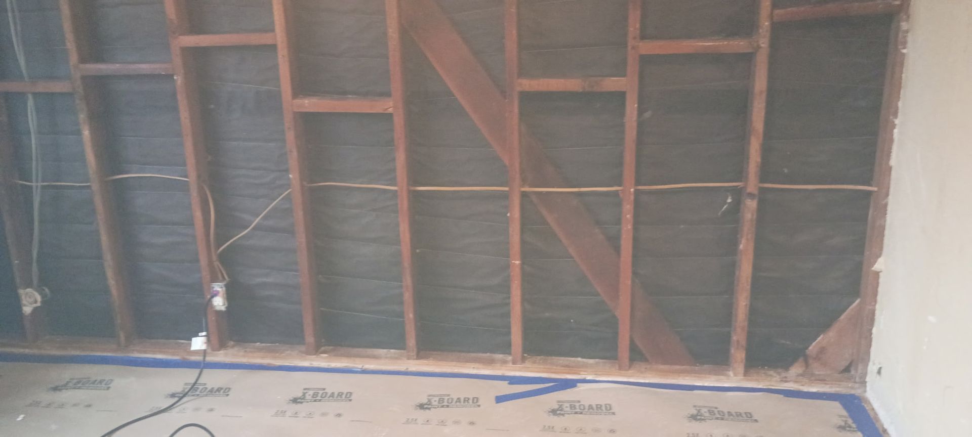 Wall framing with dark backing. Electrical wiring present. Bottom has floor covering with blue tape.