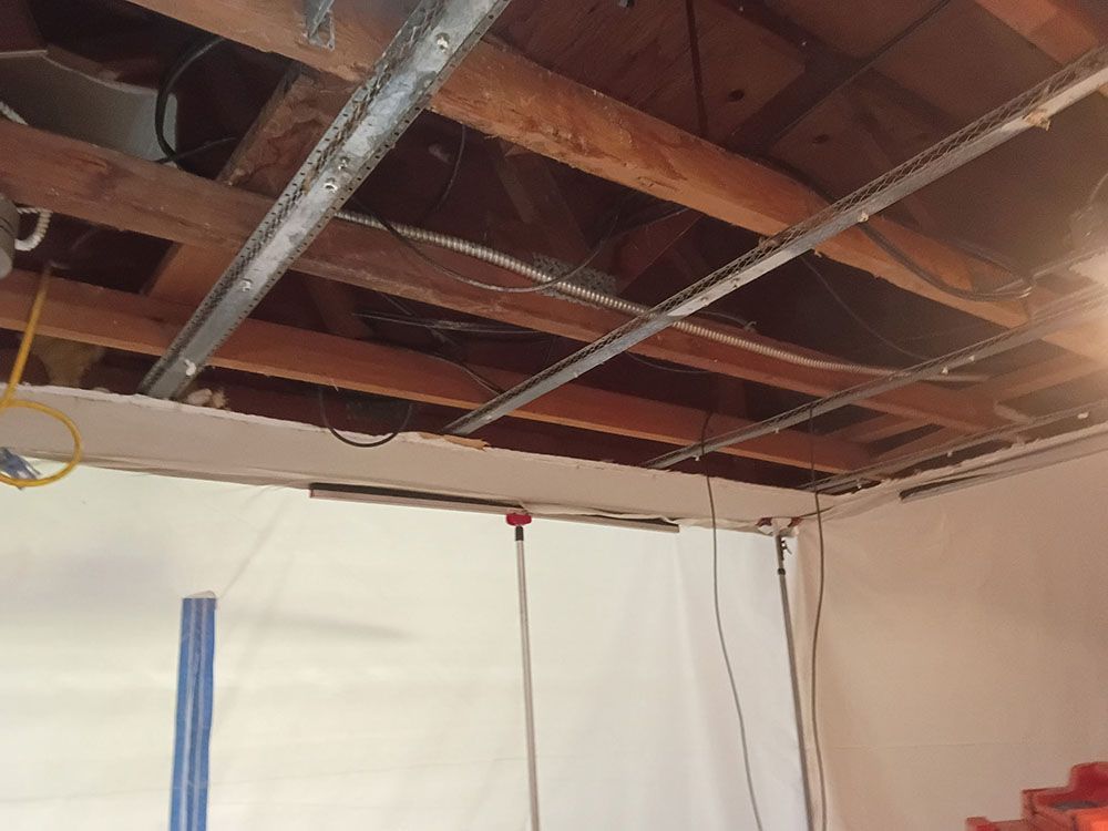 Exposed basement ceiling with wooden joists, pipes, wires, and a white wall below.