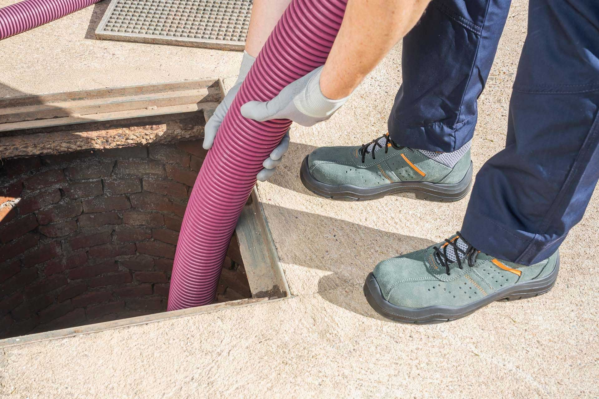 Person in work boots holding pink hose over open underground access.
