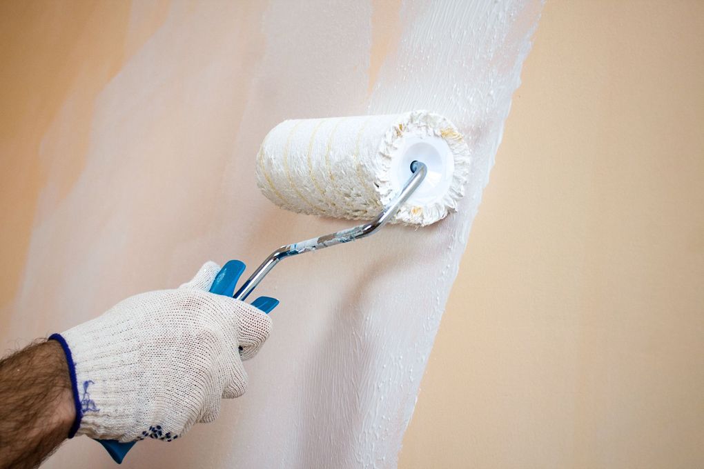Two workers in blue overalls and a maroon shirt paint a white wall with yellow and blue paint using rollers and a ladder.