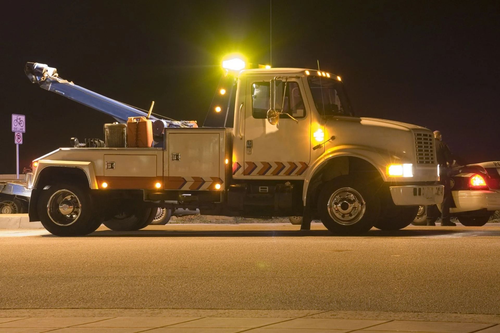 Tow truck, white, illuminated, towing a car at night under a street light.