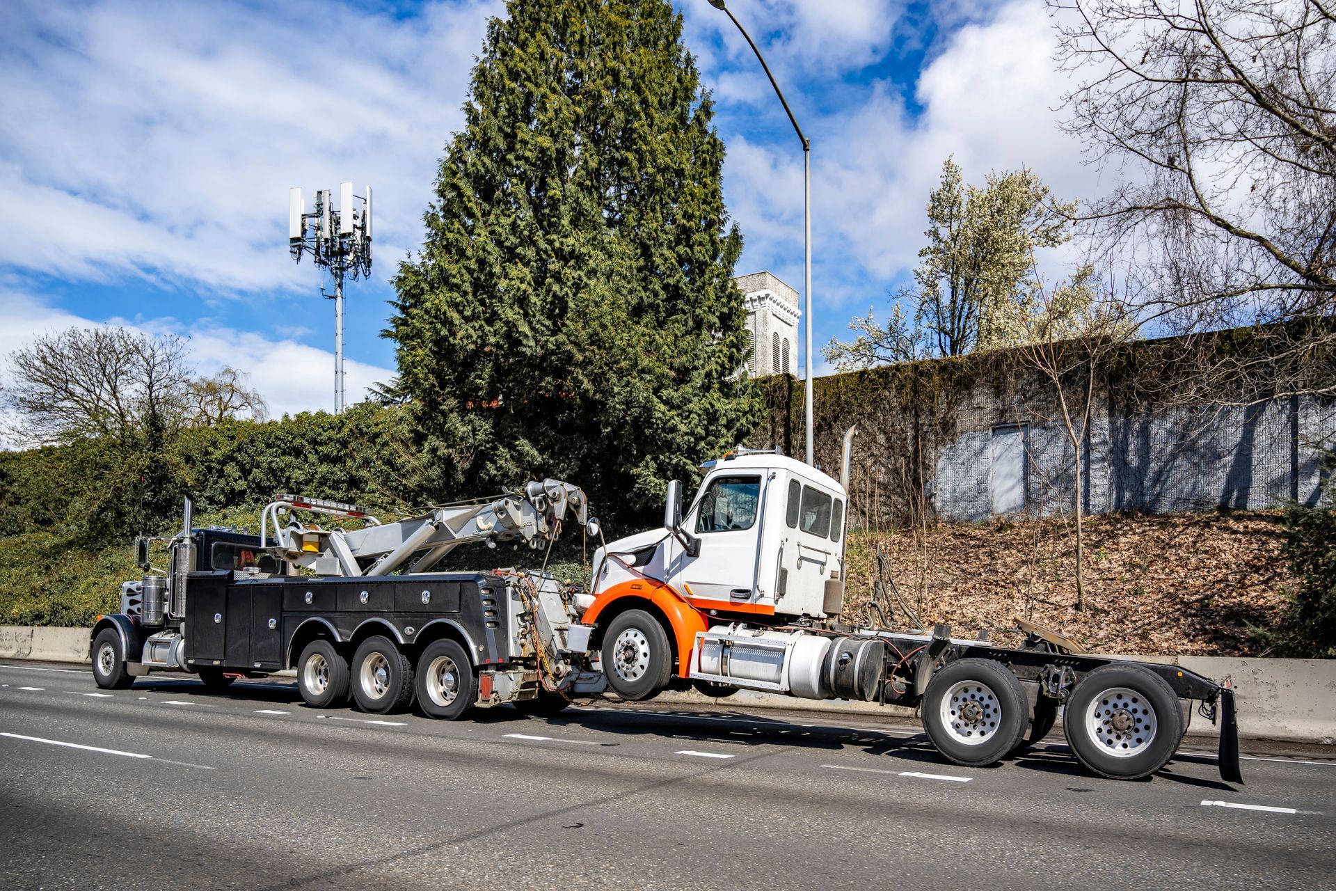 Tow truck hauling a white and orange semi-truck on a road, with trees and a building in the background.