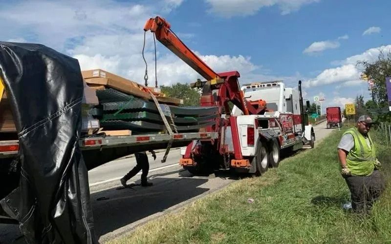 Tow truck loading cargo from a semi-trailer on the side of a highway. A worker is assisting.