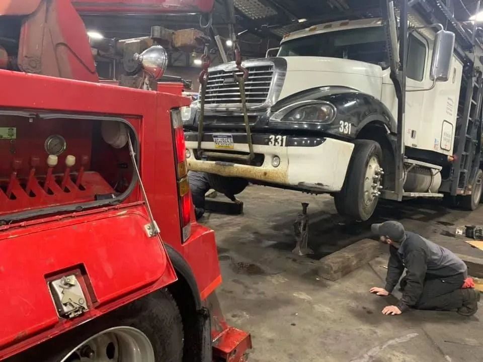 A tow truck lifting a large white truck in a repair shop; a mechanic inspects the underside.