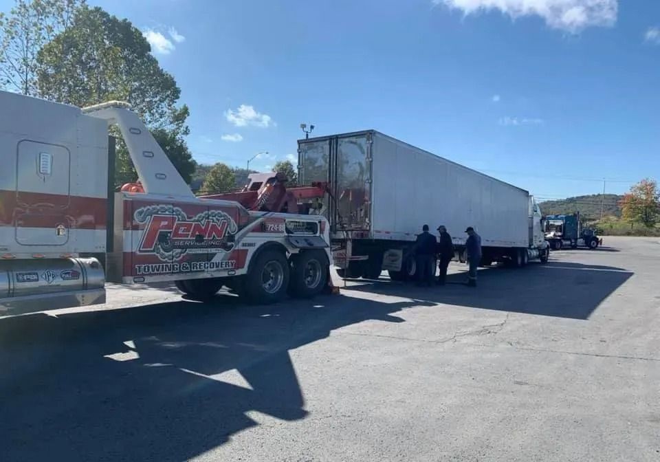 Tow truck towing a semi-truck trailer in a parking lot on a sunny day. People stand near the trailer.