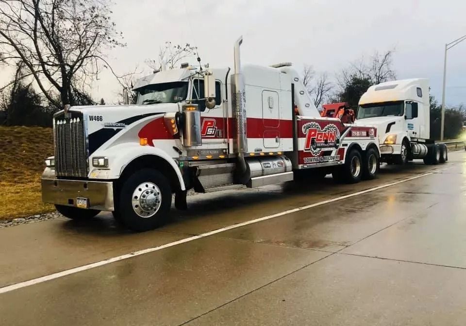 Tow truck towing a semi-truck on a wet road. Truck is white with red and black accents. Overcast day.
