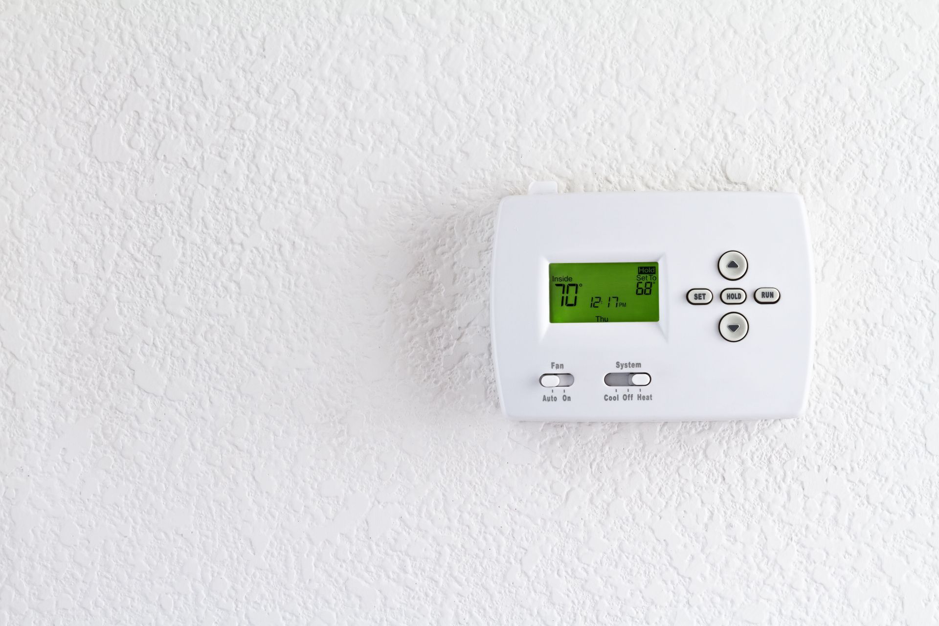 A white digital thermostat mounted on a textured white wall, displaying a room temperature of 67 degrees.