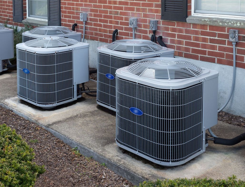 Three grey outdoor air conditioning units situated on concrete pads against a brick exterior wall of a building.