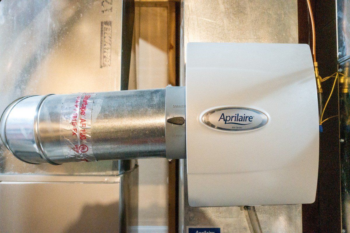 An Aprilaire whole-home humidifier unit mounted to a metallic HVAC duct system.