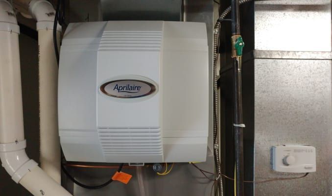 A white Aprilaire whole-home humidifier mounted on a furnace duct next to a control dial.