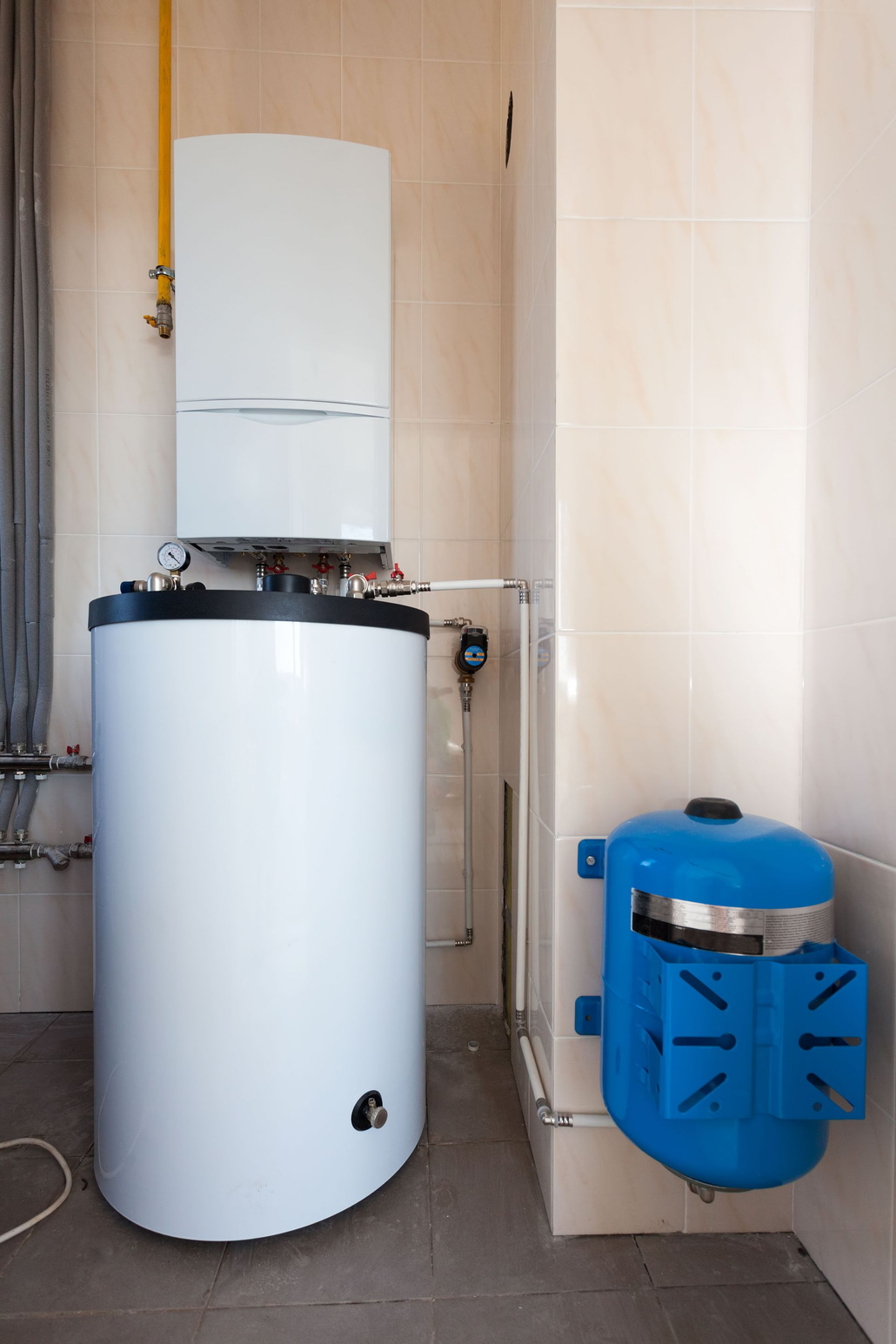 A white gas boiler and hot water cylinder sit beside a blue expansion tank on a tiled floor against a tiled wall.