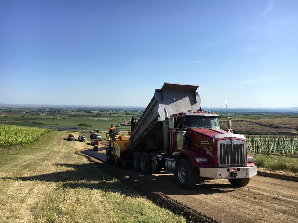 A dump truck pours asphalt into a paving machine on a rural road; fields and blue sky in the background.